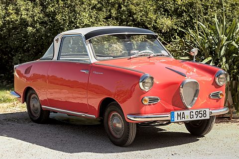Red coupé with a white roof on gravel in sunshine, seen from the front left, palms behind. Opens a larger view