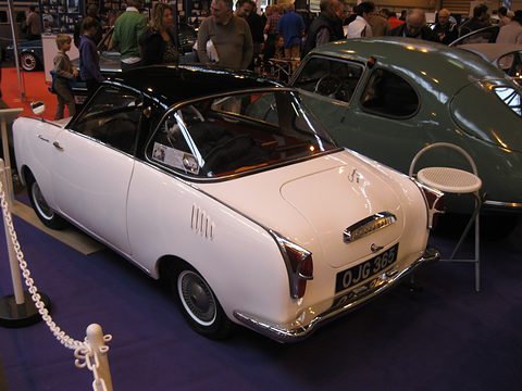 Rear three-quarter of a cream coupé with a black roof behind a chain barrier. Opens a larger view