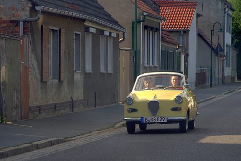 Yellow coupé with a dark grille coming towards the camera along a narrow street of old houses. Opens a larger view