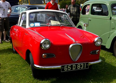 Red coupé with a white roof on grass, seen from the front left, people and a green van behind. Opens a larger view