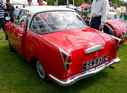Rear three-quarter of a red coupé with a white roof on grass, spectators behind. Opens a larger view