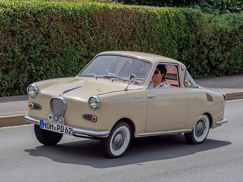 Beige coupé with whitewall tyres driving past a hedge, the driver visible. Opens a larger view