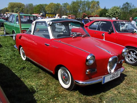 Red coupé with a white roof on grass among other classic cars, seen from the front right. Opens a larger view