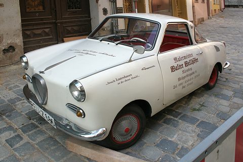 White coupé with Gothic signwriting on its door, parked on cobbles beside an old building. Opens a larger view