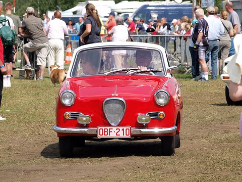 Red coupé seen from the front on grass, spectators and tents behind. Opens a larger view