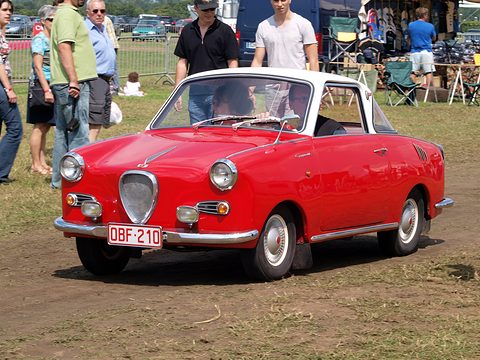 Red coupé with a white roof on grass, seen from the front left, crowd behind. Opens a larger view