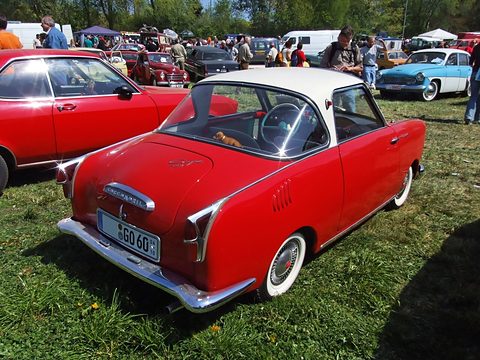 Rear view of a red coupé with a white roof on grass, spectators and tents behind. Opens a larger view
