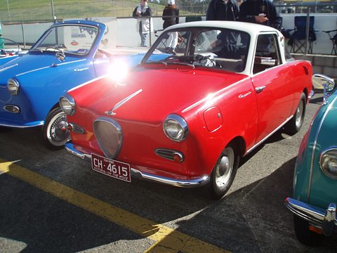 Red coupé seen from the front left on a car park, a blue car beside it. Opens a larger view