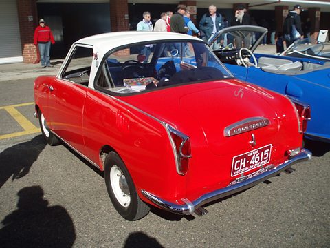 Rear of a red coupé with a white roof and club permit plate on a car park. Opens a larger view