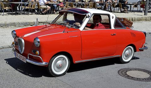 Red coupé with a white roof driving across a square, café seating behind. Opens a larger view