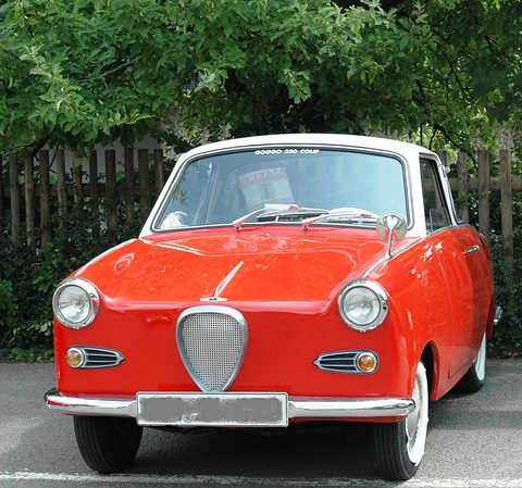 Red coupé with a white roof seen straight from the front in front of a fence and trees. Opens a larger view