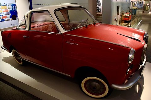 Red coupé with a white roof under museum lighting, a model railway display behind. Opens a larger view