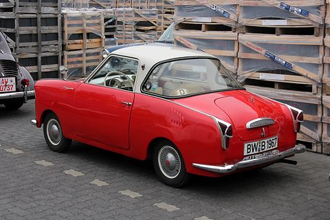 Red coupé with a white roof seen from the rear left in front of stacked wooden crates. Opens a larger view