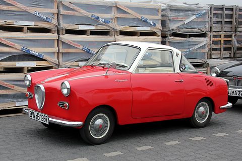 Red coupé with a white roof seen from the front left in front of stacked roofing slates. Opens a larger view