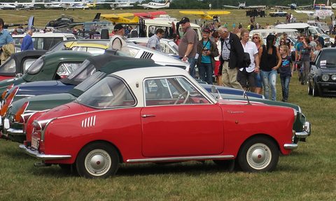 Red coupé with a white roof in side view on grass, other cars and a crowd behind. Opens a larger view