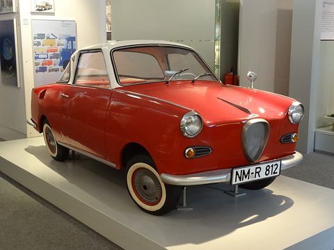 Red coupé with whitewall tyres on a white stand in an exhibition, seen from the front left. Opens a larger view