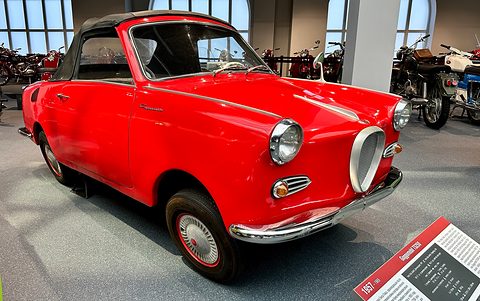 Bright red coupé with a dark roof in a museum hall with motorcycles behind. Opens a larger view