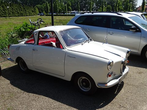 White coupé on gravel seen from the front right, modern cars parked behind. Opens a larger view