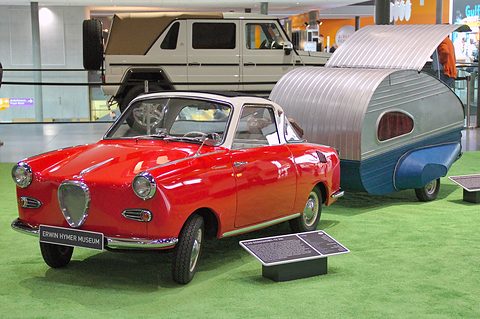 Red coupé in front of a small aluminium caravan on artificial grass at a show. Opens a larger view