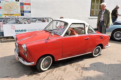 Red coupé with a white roof on paving seen from the front left, an older man standing behind. Opens a larger view