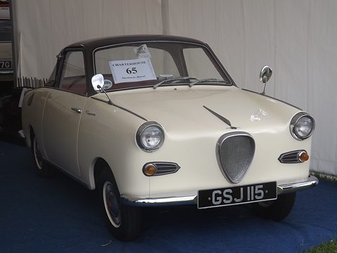 Cream coupé with a black roof on a blue carpet in a marquee, seen from the front. Opens a larger view