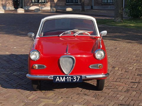 Red coupé with a white roof seen straight from the front on brick paving. Opens a larger view