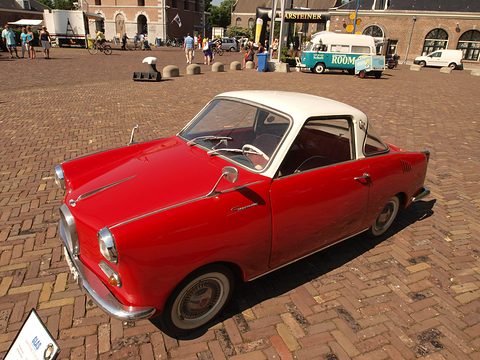 Red coupé with a white roof seen from the front left on herringbone brick paving. Opens a larger view