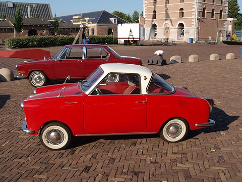Side view of a red coupé with a white roof and whitewall tyres on a brick square. Opens a larger view