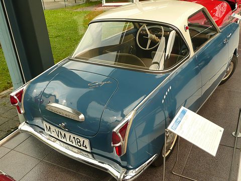 Blue coupé seen from behind with a cream roof, caption board on a stand beside it. Opens a larger view
