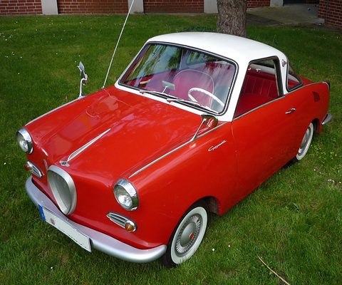 Red coupé with a white roof and whitewall tyres parked on a lawn, seen from the front left. Opens a larger view