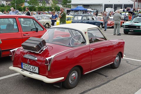 Rear of a dark red coupé with a suitcase on a rack, crowds and cars behind. Opens a larger view