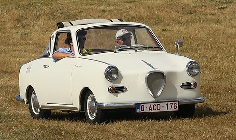 White coupé with two occupants driving across dry grass, seen from the front. Opens a larger view