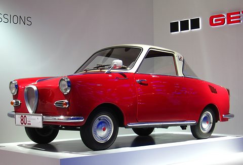 Red coupé with a white roof in side view on a white stand in a bright exhibition space. Opens a larger view