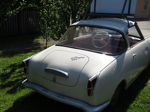Rear three-quarter of a white coupé on grass, its curved rear window and tail fins visible. Opens a larger view