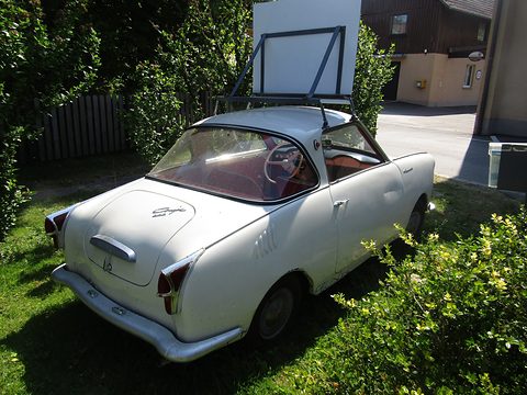 White coupé seen from the rear right beside bushes, the roof line and tail lamps visible. Opens a larger view