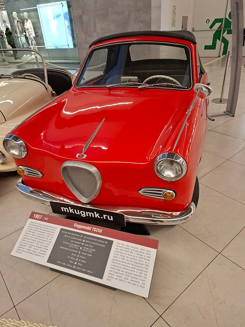 Red coupé seen from the front on a museum stand with a caption plaque. Opens a larger view