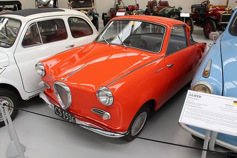Orange coupé with a white roof on a museum floor beside a white small car, caption board on the right. Opens a larger view