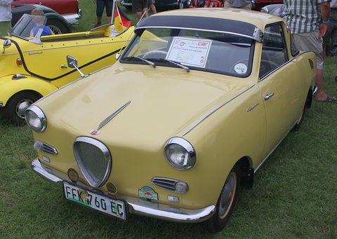 Cream coupé with a dark roof and shield-shaped grille on grass, seen from the front left. Opens a larger view