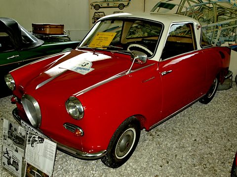 Red coupé with a white roof beside a display of old photographs and a Goggomobil sign. Opens a larger view
