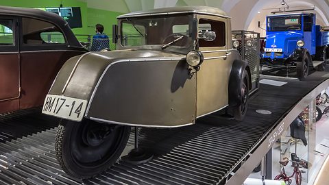 A greenish-grey three-wheeler with a pointed nose over a single front wheel on a slatted display ramp. Opens a larger view