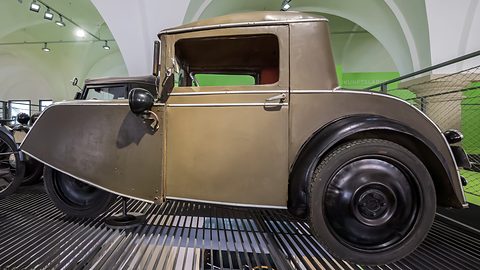 Side view of a small three-wheeler with an open door, a pointed nose and black disc wheels under vaulting. Opens a larger view