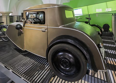 A greenish-grey two-seat three-wheeler with a short cab and a large rear wheel on a slatted ramp. Opens a larger view