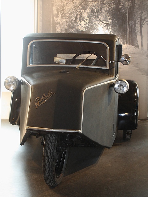 Front view of a dark grey three-wheeler with Goliath script on its pointed nose and two side lamps. Opens a larger view
