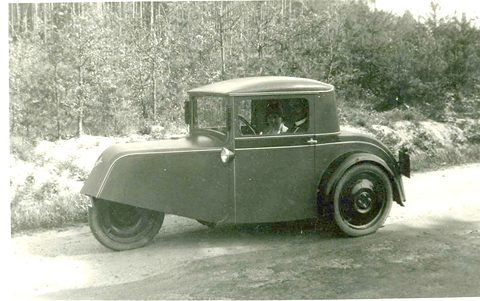 A sepia photograph of a small closed three-wheeler with a single rear wheel and rounded tail by a wooded bank.