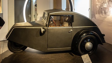 Side view of a grey three-wheeler with a sloping fastback roof and black wheel spats on a dark plinth. Opens a larger view