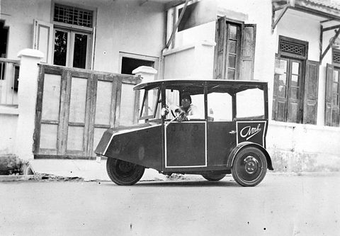 A black-and-white side view of a small open three-wheeler with a canopy roof and script on its door, a driver aboard. Opens a larger view