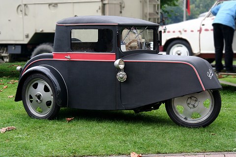 Side view of a black three-wheeled coupé with a red waist stripe, rounded tail and disc wheels on grass.
