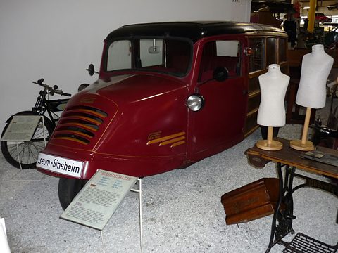 A maroon three-wheeler with a louvred rounded nose, a black roof and wood-framed side windows beside tailor’s dummies. Opens a larger view