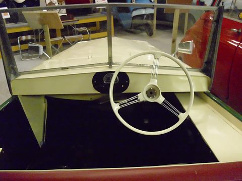Open cream cockpit with a white steering wheel and a flat windscreen, museum cars behind. Opens a larger view