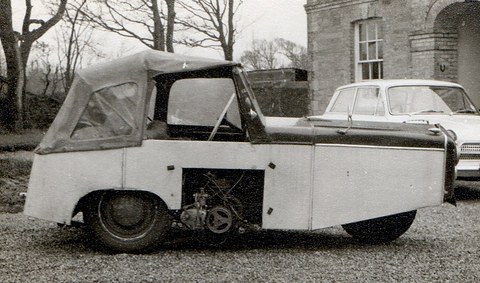 A black-and-white side view of a small boxy car with a fabric hood and a side-mounted engine by a stone house. Opens a larger view